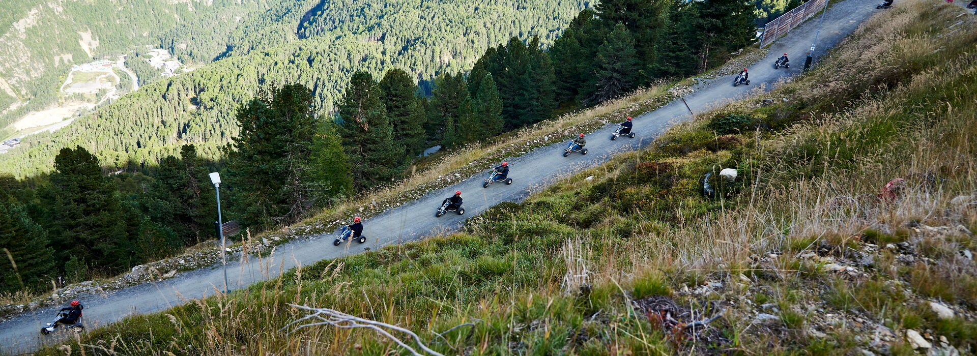 Incentive Sölden Weitwinkelaufnahme von Personen, die mit Mountaincarts von einem Berg fahren, mit Wald und Alpenpanorama