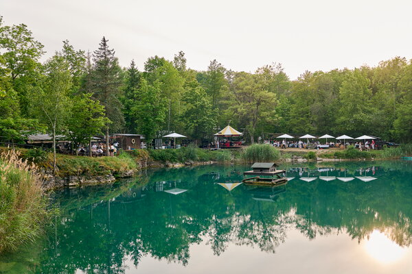 Seefest Malerischer türkisfarbener See umgeben von Bäumen, mit einem belebten Uferbereich und Sitzgelegenheiten unter Sonnenschirmen.