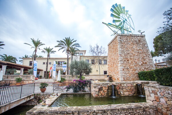Kick-off USU Mallorca mit Ein malerischer Außenbereich mit einem Steingebäude, Palmen, Wasserspiel und Windrad im Hintergrund