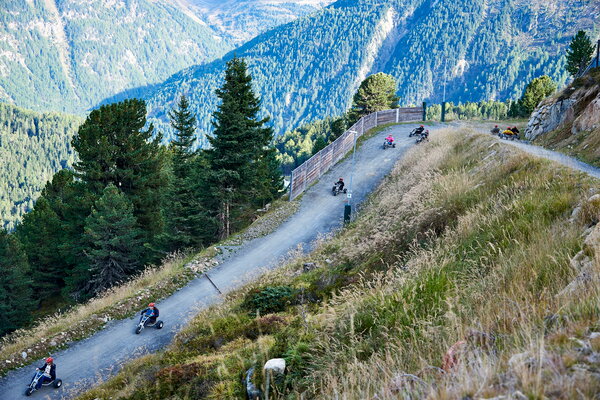 Incentive Sölden Weitwinkelaufnahme von Personen, die mit Mountaincarts auf einem Bergpfad fahren, mit Wald und Alpenpanorama